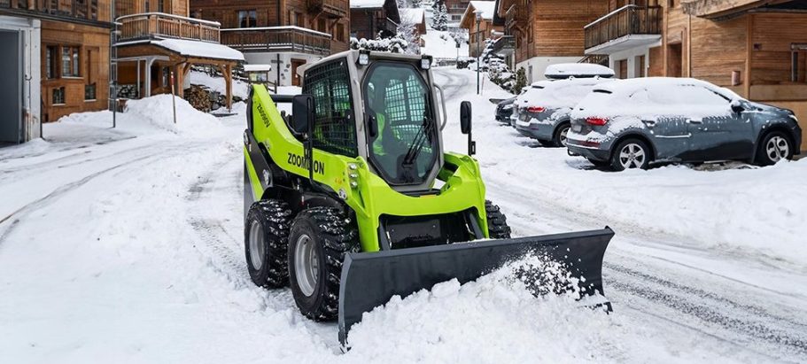 Une chargeuse compacte verte avec une lame, sur une route enneigée.